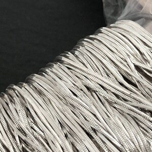 May include: A close-up of a spool of silver-colored chain. The chain is made of small, flat, interlocking links, creating a textured surface. The chain is tightly wound, suggesting it is new and unused. The background is dark.