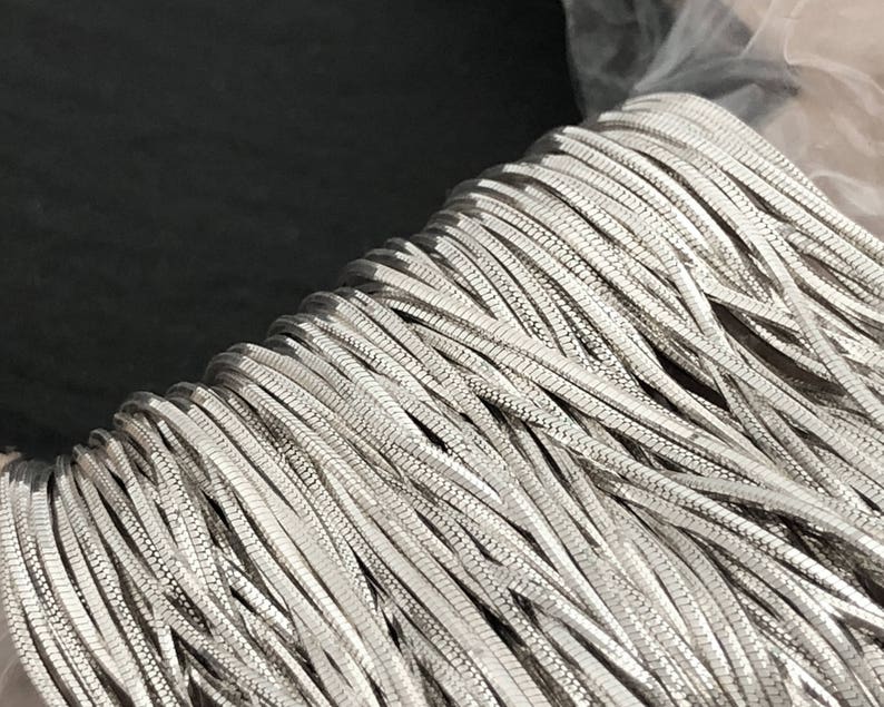 May include: A close-up of a spool of silver-colored chain. The chain is made of small, flat, interlocking links, creating a textured surface. The chain is tightly wound, suggesting it is new and unused. The background is dark.