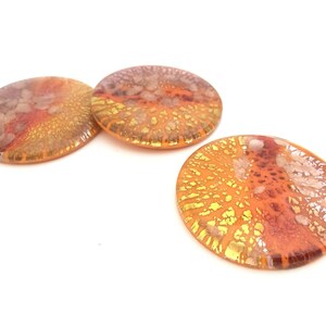 May include: Three round, glass decorative pieces with a blend of orange, gold, and reddish-brown hues. The glass has a textured, speckled appearance, with a lighter, almost iridescent quality in some areas. The pieces are set against a white background.