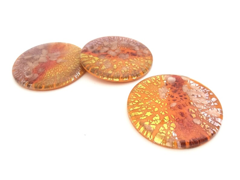 May include: Three round, glass decorative pieces with a blend of orange, gold, and reddish-brown hues. The glass has a textured, speckled appearance, with a lighter, almost iridescent quality in some areas. The pieces are set against a white background.