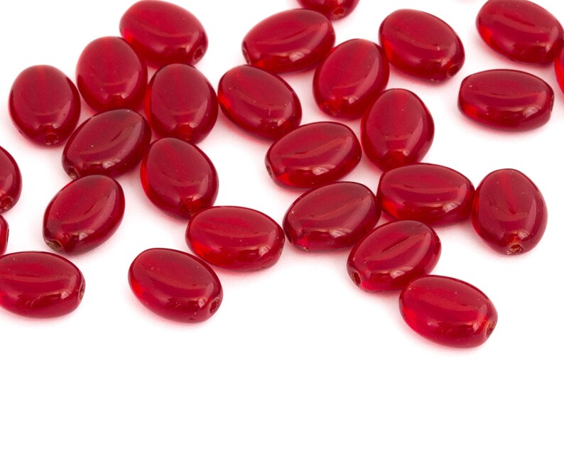 May include: A collection of red glass oval beads, scattered on a white surface.