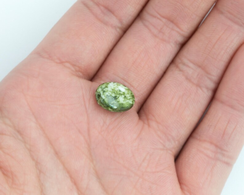 May include: An oval-shaped, green and white gemstone bead. The bead has a speckled pattern and a polished surface. The bead is held in an open hand against a white background. The bead is approximately 0.5 inches in length.