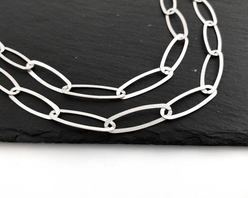 Puede incluir: Un collar de cadena plateada con eslabones ovalados grandes. El collar se muestra sobre una superficie negra, con los eslabones dispuestos en un patr&oacute;n fluido. La cadena es de metal brillante.