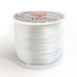 May include: A spool of clear, white elastic beading cord. The spool has a white base and a label with Japanese text and a diamond graphic. The cord is tightly wound, showing a smooth, reflective surface.