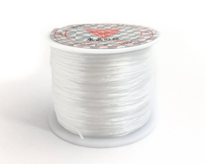 May include: A spool of clear, white elastic beading cord. The spool has a white base and a label with Japanese text and a diamond graphic. The cord is tightly wound, showing a smooth, reflective surface.
