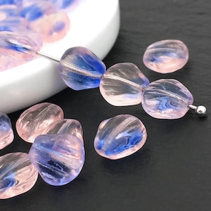 Puede incluir: Cuentas de vidrio translúcidas rosas y azules con un diseño en espiral. Las cuentas están ensartadas en un alambre plateado y están dispersas sobre una superficie oscura. Las cuentas tienen una forma redondeada e irregular, y están parcialmente dentro de un cuenco blanco.