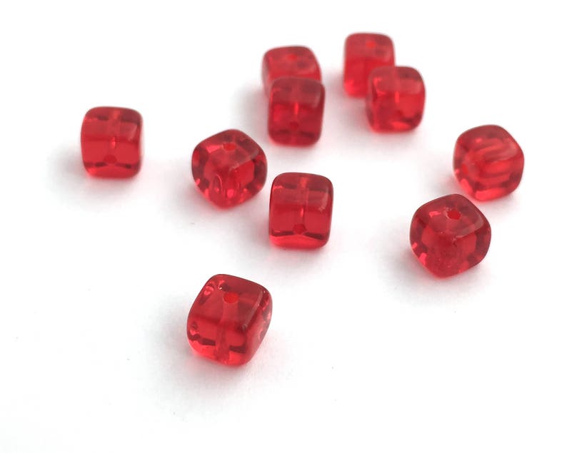 Puede incluir: Un conjunto de cuentas rojas transl&uacute;cidas en forma de cubo con bordes redondeados. Cada cuenta tiene un peque&ntilde;o agujero para ensartar. Las cuentas est&aacute;n dispersas sobre una superficie blanca, mostrando su textura brillante y su color vibrante. Ideal para la creaci&oacute;n de joyas.