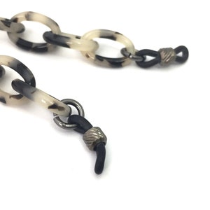May include: A black and white tortoiseshell patterned plastic chain with black rubber ends and silver metal accents. The chain is designed to hold glasses.