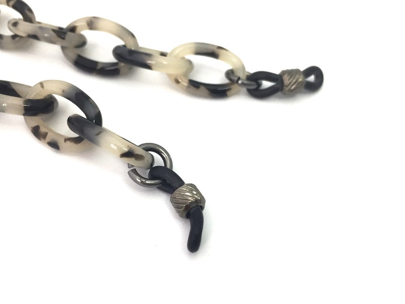 May include: A black and white tortoiseshell patterned plastic chain with black rubber ends and silver metal accents. The chain is designed to hold glasses.
