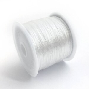 May include: A spool of clear, elastic beading cord. The cord is tightly wound around a white plastic spool. The image is a close-up shot against a white background, highlighting the texture and transparency of the cord.