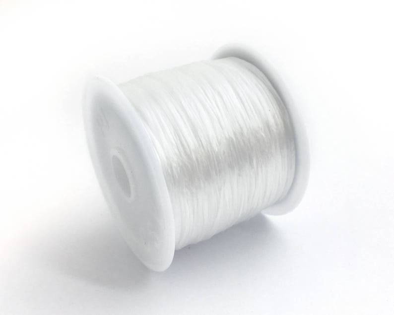 May include: A spool of clear, elastic beading cord. The cord is tightly wound around a white plastic spool. The image is a close-up shot against a white background, highlighting the texture and transparency of the cord.
