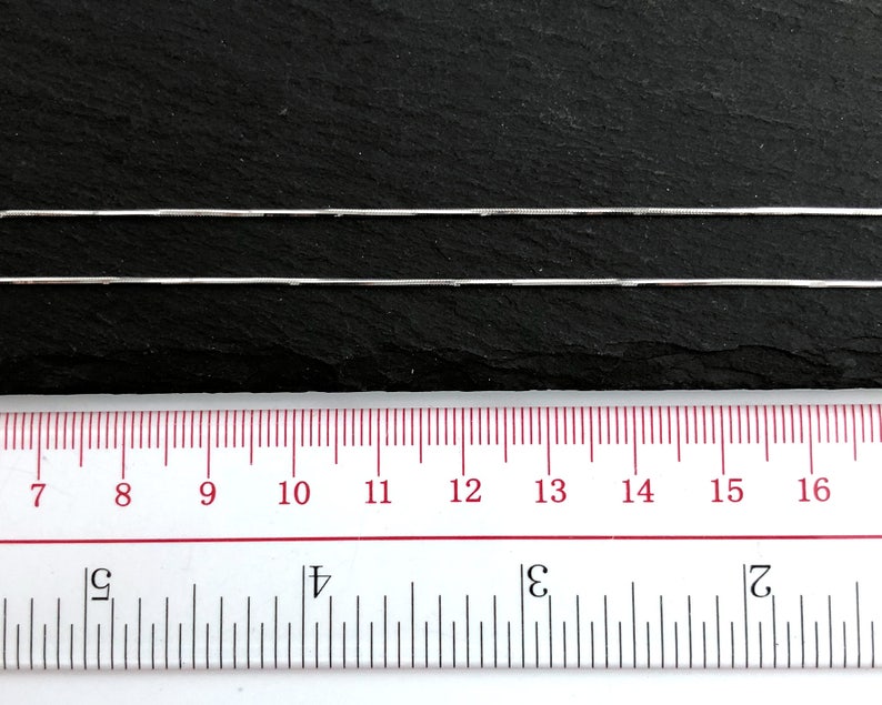 May include: Two silver-colored snake chains are displayed against a dark background. A ruler with inch markings is positioned below the chains, showing measurements from 7 to 16 inches. The chains are thin and reflective.