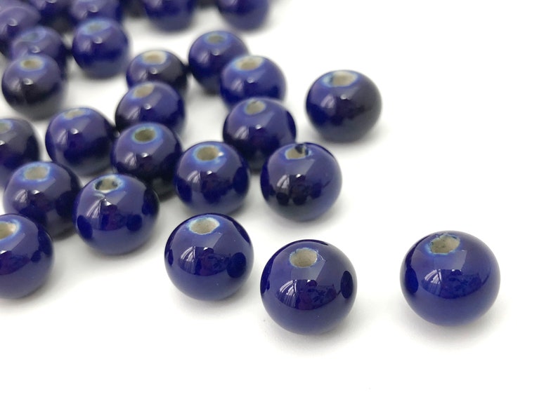 May include: A collection of glossy, dark blue ceramic beads. Each bead is round with a small hole and a lighter blue-grey area around the hole. The beads are scattered on a white surface.