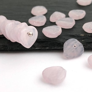 Puede incluir: Una colección de cuentas de cuarzo rosa en forma de corazón de color rosa pálido. Las cuentas están esparcidas sobre una superficie de pizarra negra.