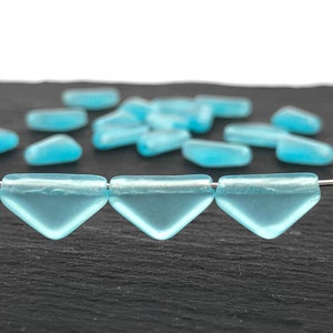May include: Three light blue glass triangle beads strung on a silver wire. The beads are translucent and have a frosted finish. The beads are on a black surface with other beads scattered in the background.