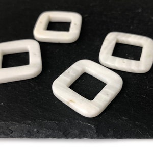Peut inclure: Quatre perles carr&eacute;es blanches avec un trou au centre. Les perles ont un motif marbr&eacute; et sont sur une surface noire.