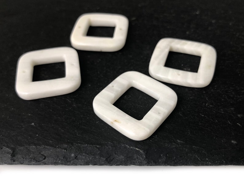 Peut inclure: Quatre perles carr&eacute;es blanches avec un trou au centre. Les perles ont un motif marbr&eacute; et sont sur une surface noire.