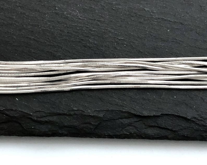 May include: Close-up of several strands of silver-colored snake chain. The chains are layered and slightly curved, creating a textured appearance. The background is a dark, textured surface.