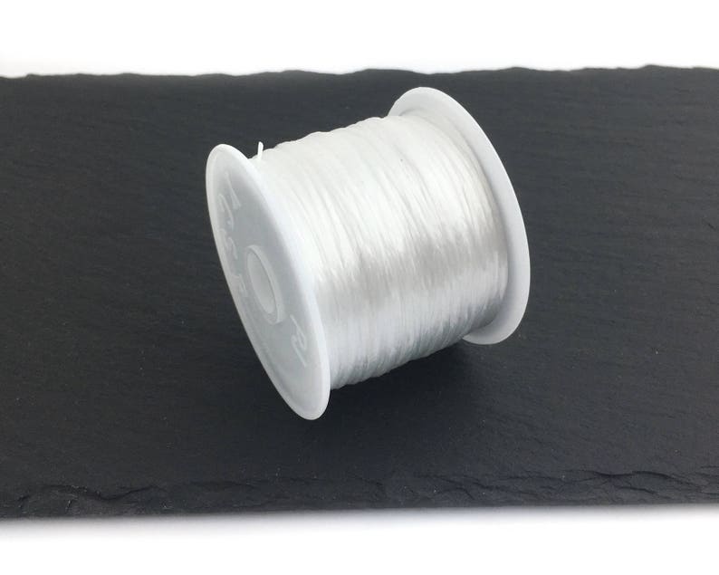 May include: A spool of clear, elastic beading cord. The cord is tightly wound on a white plastic spool. The spool is resting on a dark, textured surface. This cord is ideal for jewelry making and crafting projects.