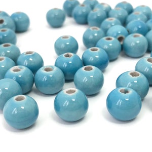 May include: A collection of light blue ceramic beads with a glossy finish. Each bead is round with a hole running through the center. The beads are scattered on a white surface, ideal for jewelry making and crafts.