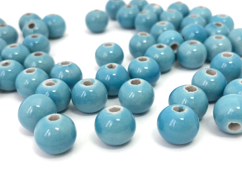 May include: A collection of light blue ceramic beads with a glossy finish. Each bead is round with a hole running through the center. The beads are scattered on a white surface, ideal for jewelry making and crafts.