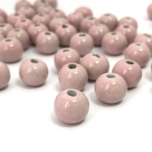 May include: A collection of pale pink ceramic beads. Each bead is round with a small hole at the top. The beads have a glossy finish and are scattered on a white surface, creating a soft, delicate aesthetic.
