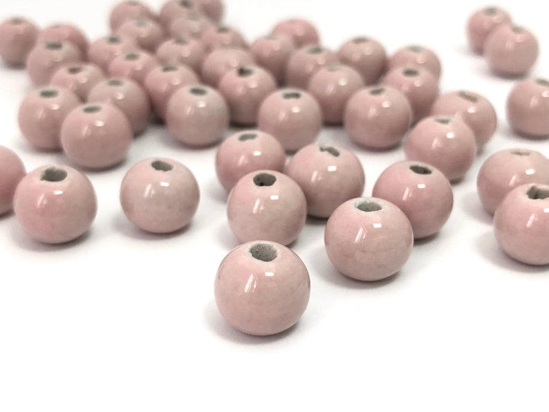 May include: A collection of pale pink ceramic beads. Each bead is round with a small hole at the top. The beads have a glossy finish and are scattered on a white surface, creating a soft, delicate aesthetic.