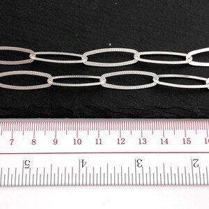 Puede incluir: Primer plano de dos hebras de cadena de eslabones ovalados plateados. Los eslabones tienen una superficie texturizada. Una regla con medidas en cent&iacute;metros est&aacute; en primer plano, mostrando la longitud de la cadena. El fondo es oscuro.