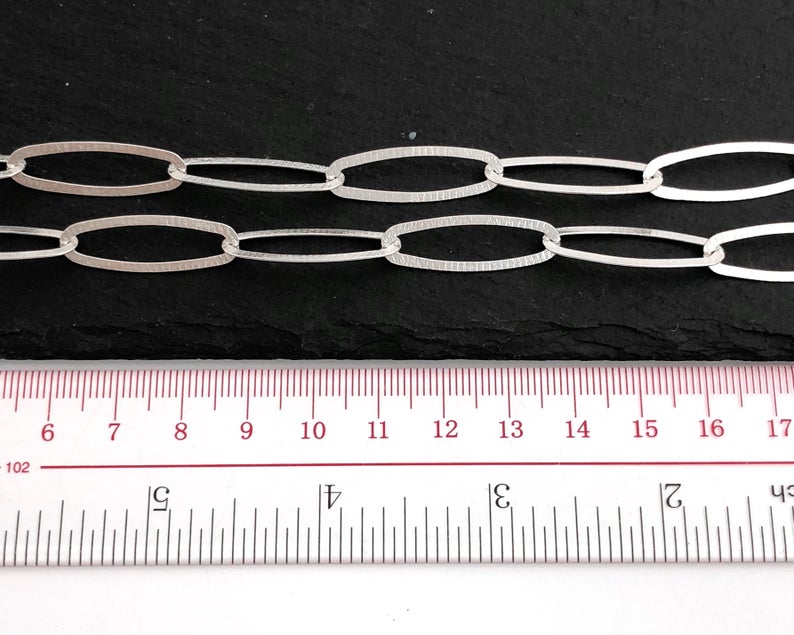 Puede incluir: Primer plano de dos hebras de cadena de eslabones ovalados plateados. Los eslabones tienen una superficie texturizada. Una regla con medidas en cent&iacute;metros est&aacute; en primer plano, mostrando la longitud de la cadena. El fondo es oscuro.