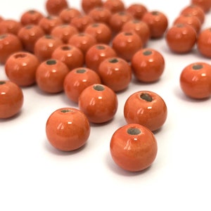 May include: A collection of orange-red ceramic beads with a glossy finish. Each bead is spherical with a small hole running through the center. The beads are scattered on a white surface, suitable for jewelry making or crafts.