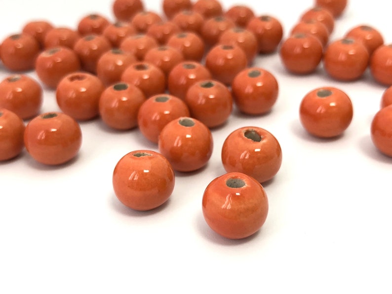 May include: A collection of orange-red ceramic beads with a glossy finish. Each bead is spherical with a small hole running through the center. The beads are scattered on a white surface, suitable for jewelry making or crafts.