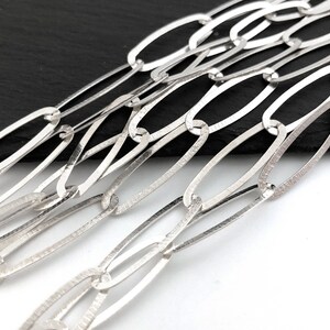 Puede incluir: Primer plano de un collar de cadena plateada con eslabones ovalados alargados. Los eslabones tienen un aspecto texturizado y martillado, y la cadena se muestra sobre un fondo blanco y negro. La joyer&iacute;a es probablemente hecha a mano.
