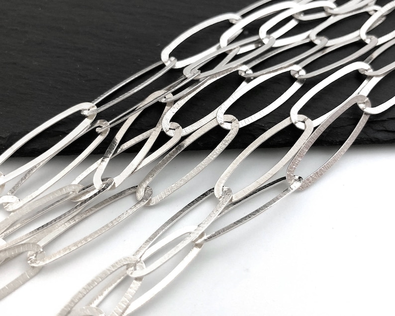 Puede incluir: Primer plano de un collar de cadena plateada con eslabones ovalados alargados. Los eslabones tienen un aspecto texturizado y martillado, y la cadena se muestra sobre un fondo blanco y negro. La joyer&iacute;a es probablemente hecha a mano.