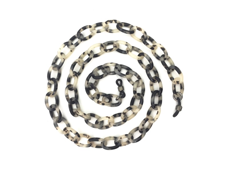 May include: A black and white tortoiseshell patterned plastic chain for glasses. The chain is made of oval links and has metal clasps on each end.