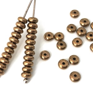 以下が含まれることがあります： 小さな丸い金色のビーズの山をクローズアップした写真です。ビーズは白い表面に緩く積み重ねられています。画像の左側にはビーズの糸が2本あり、それぞれ銀色のワイヤーに通されています。