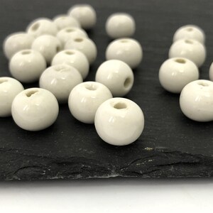 May include: A collection of off-white, spherical beads with a glossy finish. Each bead has a hole running through the center, suggesting they are for stringing. The beads are scattered on a dark, textured surface, likely for crafting or jewelry making.