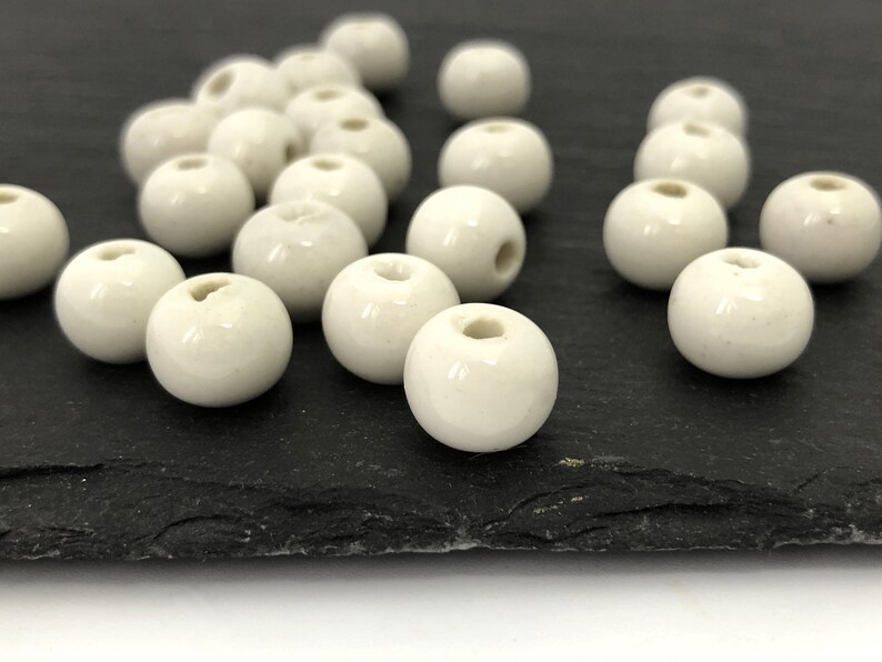 May include: A collection of off-white, spherical beads with a glossy finish. Each bead has a hole running through the center, suggesting they are for stringing. The beads are scattered on a dark, textured surface, likely for crafting or jewelry making.