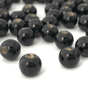 May include: A collection of glossy black round beads with a small, light-colored hole at the top. The beads are scattered across a white surface, creating a visually appealing arrangement. These beads are suitable for jewelry making.