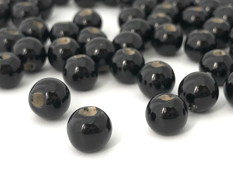May include: A collection of glossy black round beads with a small, light-colored hole at the top. The beads are scattered across a white surface, creating a visually appealing arrangement. These beads are suitable for jewelry making.