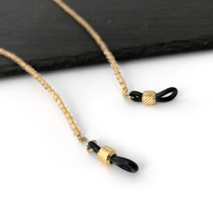 May include: A gold beaded eyeglass chain with black silicone ends. The chain is made of small, round beads and has a simple, elegant design.