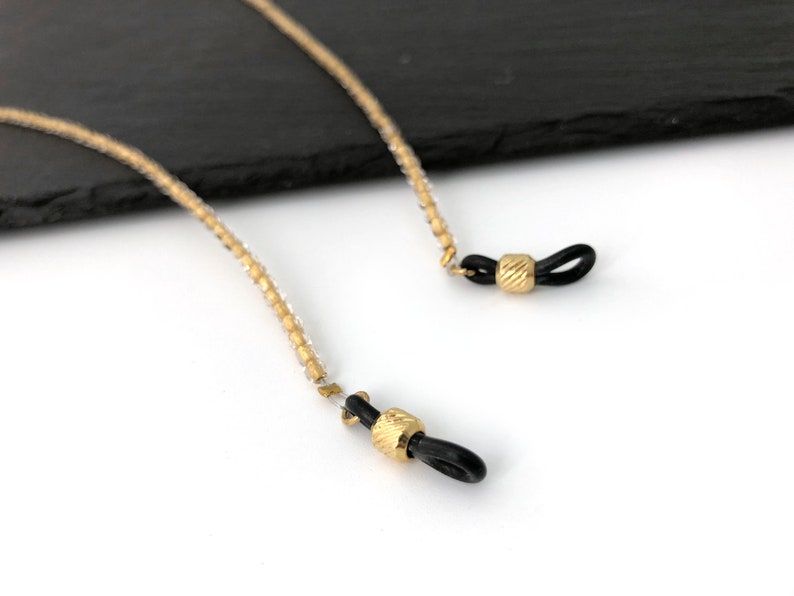 May include: A gold beaded eyeglass chain with black silicone ends. The chain is made of small, round beads and has a simple, elegant design.