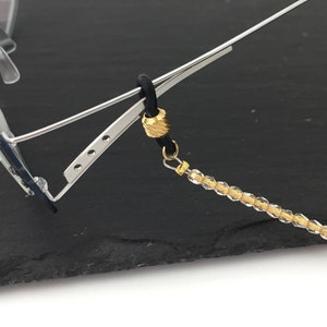 May include: A gold-toned beaded eyeglass chain with a black rubber cord and a gold-toned bead. The chain is attached to a pair of eyeglasses.