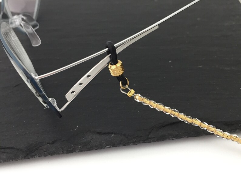 May include: A gold-toned beaded eyeglass chain with a black rubber cord and a gold-toned bead. The chain is attached to a pair of eyeglasses.