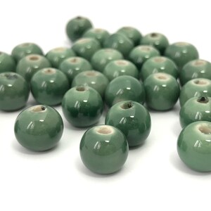 May include: A collection of glossy, round, green ceramic beads. Each bead has a small hole running through the center. The beads are scattered on a white surface, with some in focus and others slightly blurred.