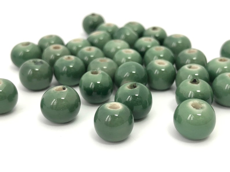 May include: A collection of glossy, round, green ceramic beads. Each bead has a small hole running through the center. The beads are scattered on a white surface, with some in focus and others slightly blurred.
