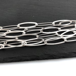 Puede incluir: Primer plano de un collar de cadena plateada con eslabones ovalados. La cadena est&aacute; hecha de un material met&aacute;lico brillante y se muestra sobre un fondo oscuro. Los eslabones est&aacute;n entrelazados, creando un patr&oacute;n texturizado.