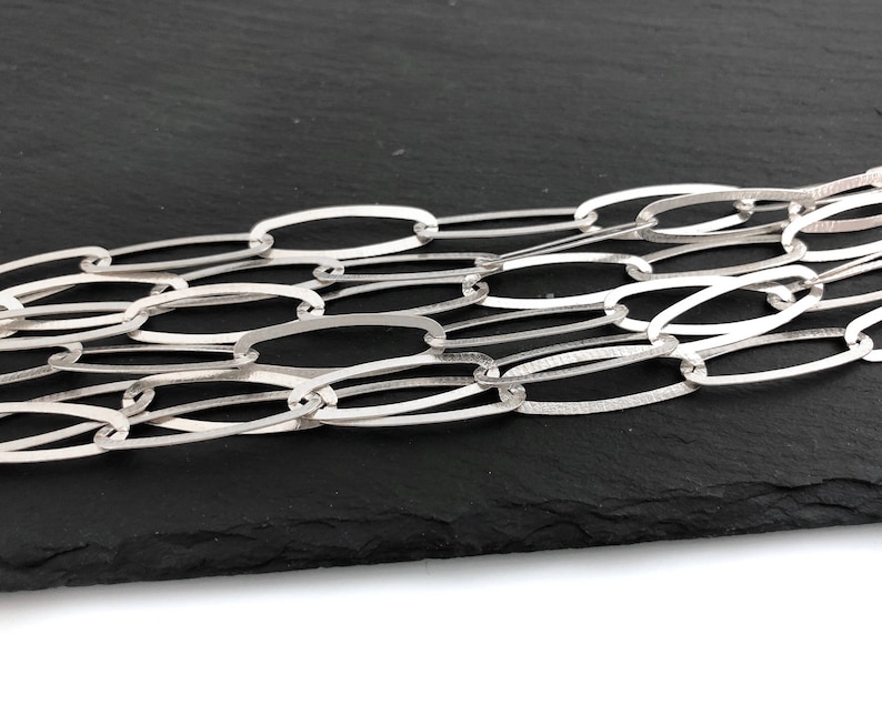 Puede incluir: Primer plano de un collar de cadena plateada con eslabones ovalados. La cadena est&aacute; hecha de un material met&aacute;lico brillante y se muestra sobre un fondo oscuro. Los eslabones est&aacute;n entrelazados, creando un patr&oacute;n texturizado.