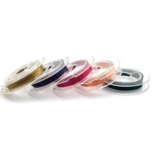May include: Six spools of wire in different colors, including gold, purple, pink, copper, green, and black. The spools are made of clear plastic and are arranged in a row.