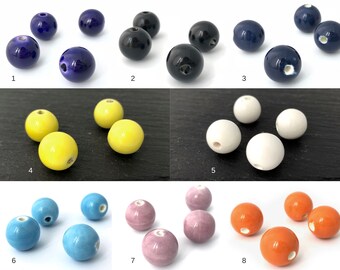 Könnte beinhalten: Eine Auswahl an Keramikperlen in verschiedenen Farben: Dunkelblau, Schwarz, Gelb, Weiß, Hellblau, Rosa und Orange. Jede runde Perle hat ein Loch zum Auffädeln. Die Perlen sind auf einer dunklen Oberfläche angeordnet.
