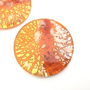 May include: Two orange and yellow dichroic glass pendants with a circular shape. The pendants have a hole at the top for stringing. The glass has a textured, layered appearance with gold and white accents. The pendants are set against a white background.